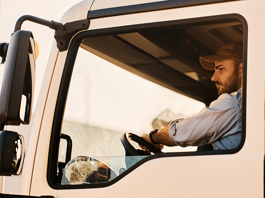 TT-BlogImage-0723-ArticleImages-5-1 Trailer Transit Inc. | A man wearing a cap is driving a truck, viewed from the passenger side window, with sunlight filtering through. The scene captures the daily life of owner-operators working for transport companies.