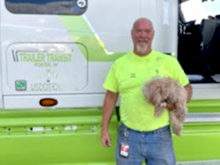 TT-BlogImage-1023-ArticleImages-1 Trailer Transit Inc. | A man wearing a neon yellow shirt stands in front of a truck, holding a small dog. The truck has “TRAILER TRANSIT” printed on it along with other text and markings, indicating it's part of an owner-operator transport company.