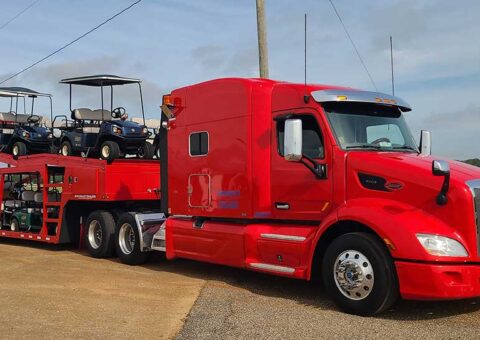 Trailer Transit Inc. | A red semi-truck engaged in power-only trailer transport, carrying a trailer loaded with golf carts parked on a paved lot under a clear sky.