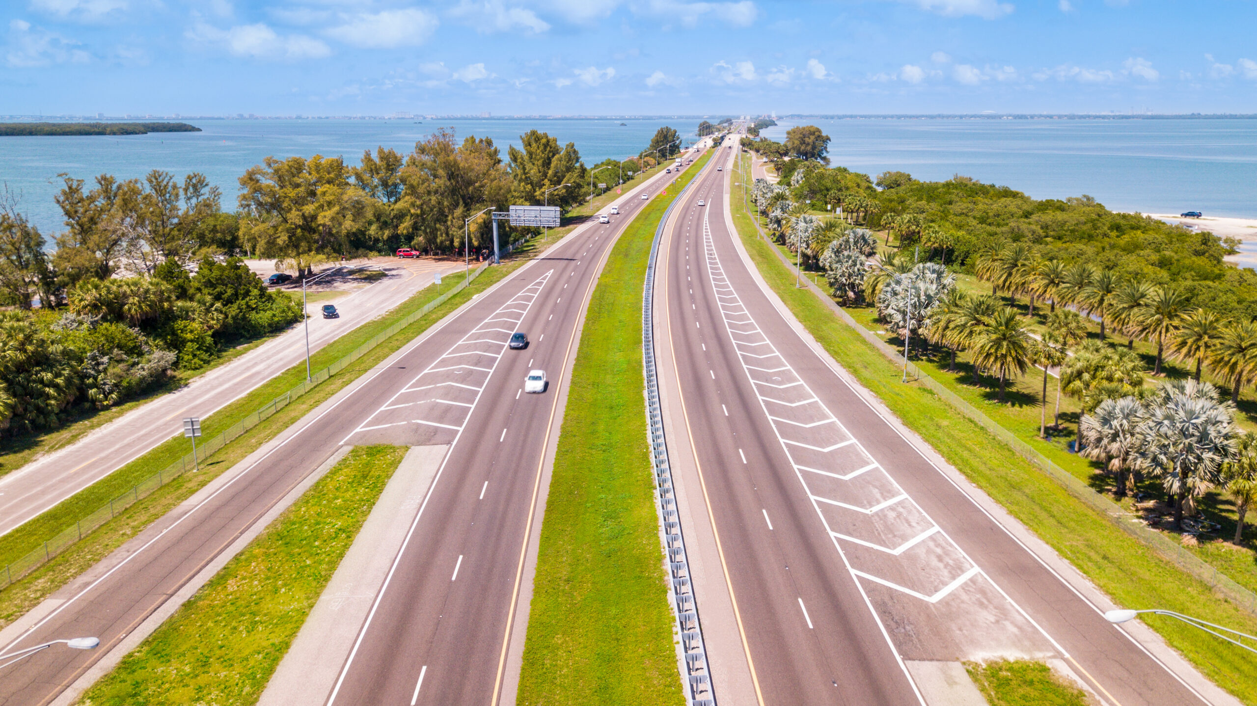 Highway or Freeway road. Low traffic on the way. Highway for any transportation. Road for cars and truck. The Best US American roads for travels. Gulf of Mexico. Wide road with many lanes. Aerial view Trailer Transit Inc. | Aerial view of a wide, divided highway with few vehicles, surrounded by greenery and water on a clear day.