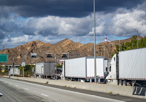 Line of big rig semi trucks with loaded semi trailers standing on the at the weighing station with scales for weighing along the axes. Trailer Transit, Inc. | semi trucks in line at US weigh station at port of entry