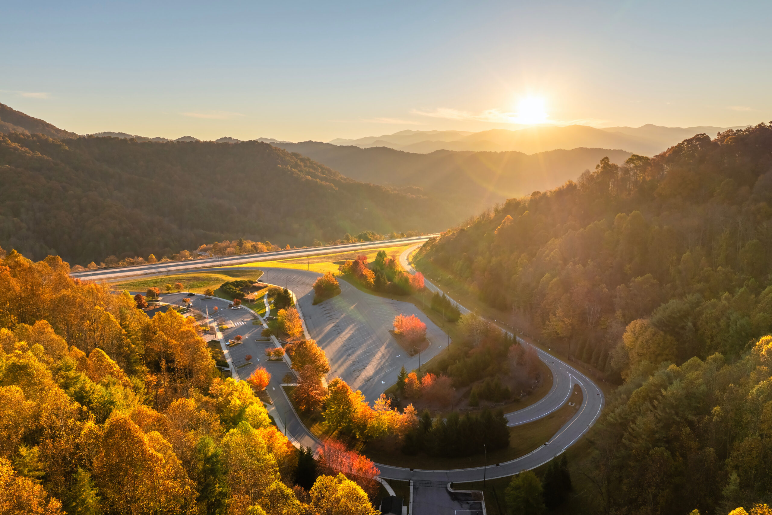 Truck stop with Beech Glen North Carolina welcome center near busy American I-26 interstate freeway with driving cars and trucks. Recreational parking during interstate travel in fall season Trailer Transit Inc. | A winding road curves through a forested mountainous area at sunset, with vibrant autumn foliage and a sunlit sky.