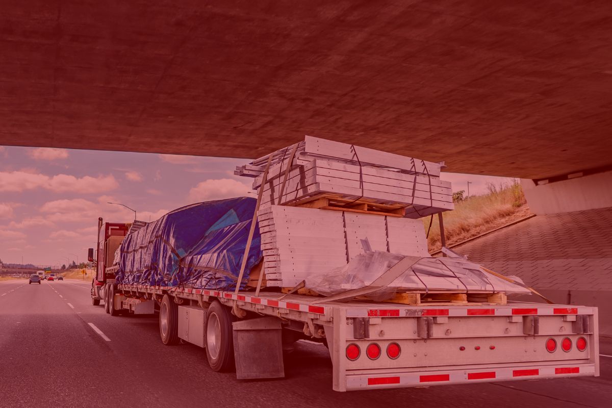 2 Trailer Transit Inc. | A flatbed truck hauled by a Power-Only driver, loaded with large, covered cargo and construction materials, drives on a highway beneath an overpass.
