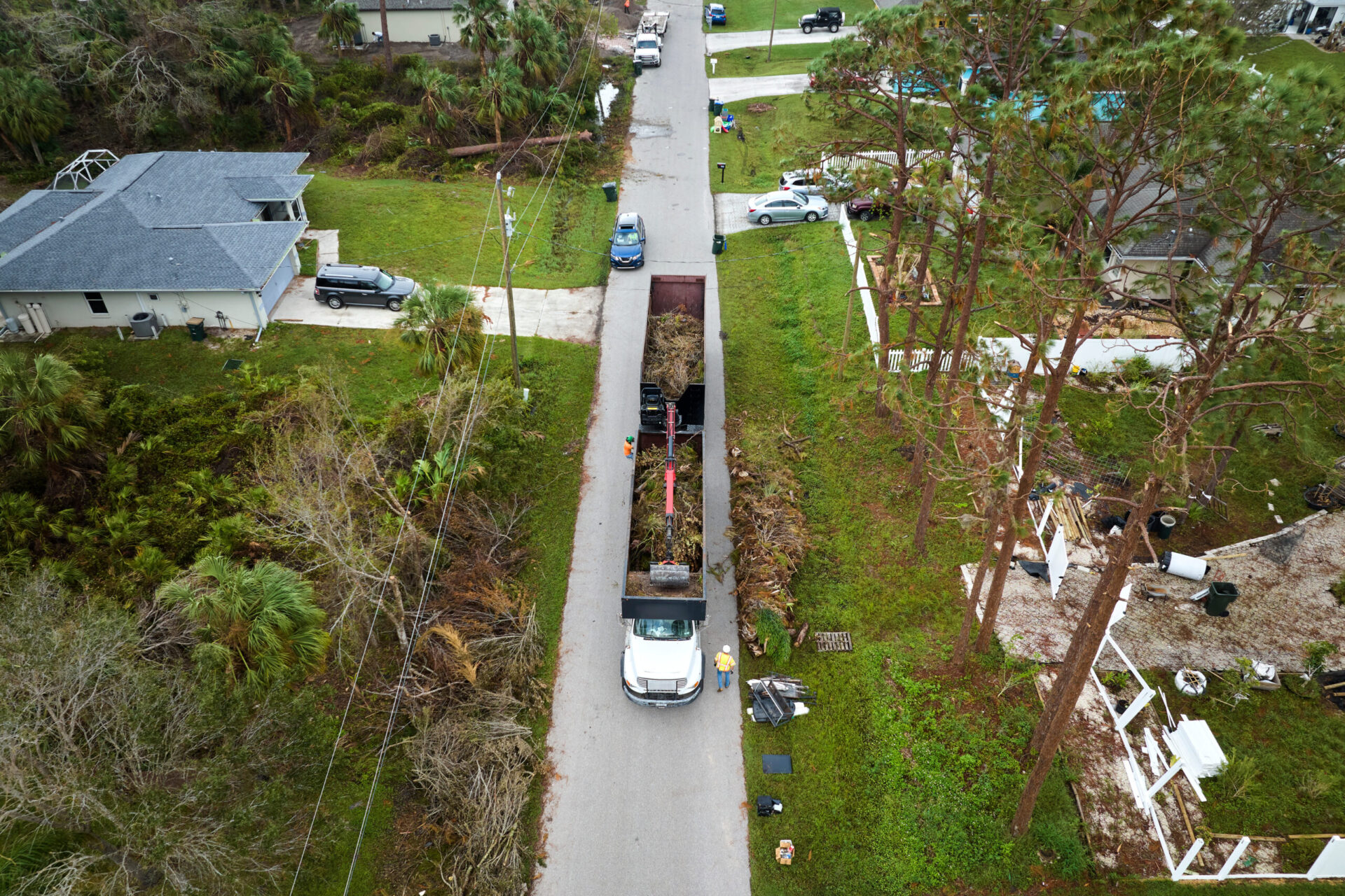 Top view of Hurricane Ian special aftermath recovery dump truck picking up tree branches debris from Florida rural streets. Dealing with consequences of natural disaster Trailer Transit Inc. | A Trucking unit with a trailer is collecting debris from a residential street lined with damaged trees and houses, likely after a storm.