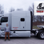 Trailer Transit Inc. | Proud owner operator, Brian, stands before his white semi-truck. A “Trailer Transit Inc” badge in the corner honors him as Owner Operator of the Month.