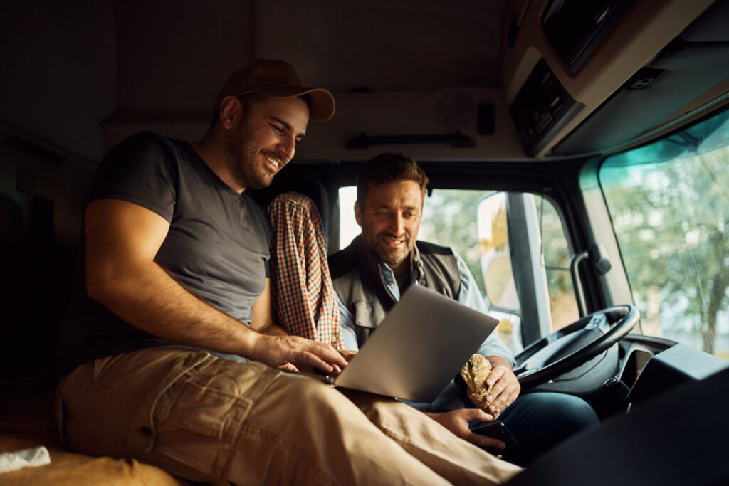Trailer Transit Inc. | Two men sitting in a truck cab, one using a laptop and the other holding a sandwich, both smiling and looking at the screen—showcasing the joy of bringing companions on the road in trucking.