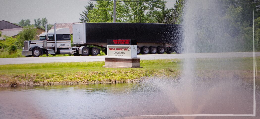 0126 Blog-6 Trailer Transit Inc. | A semi-truck offering power-only trucking with a companion drives past a company sign for Paladin Transit Inc. near a pond with a water fountain during the daytime.