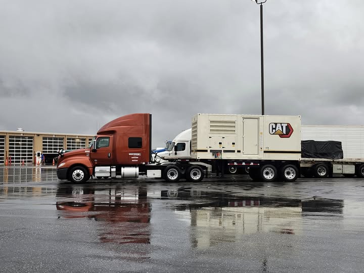 truck hauling Trailer Transit Inc. | A red semi-truck with attached trailers, including a large CAT-branded generator unit, parked on wet asphalt under a cloudy sky—an impressive sight from the best owner operator company.