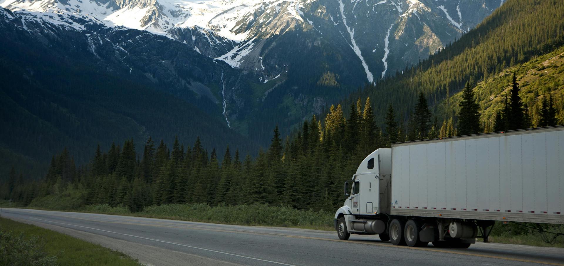 Trailer Transit, Inc. | A white semi-truck drives on a highway through a forested valley with snow-capped mountains in the background.