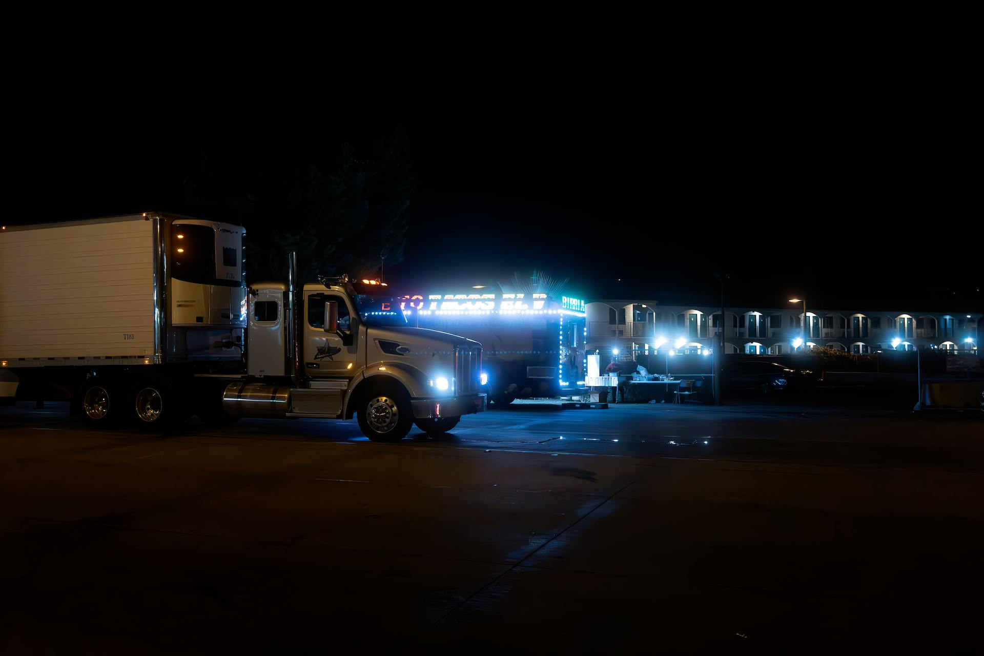 Trailer Transit, Inc. | A large semi-truck parked at night near a brightly lit building with multiple illuminated windows and a neon sign.