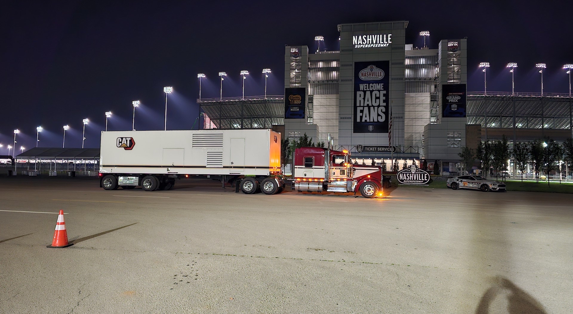 Trailer Transit, Inc. | A semi-truck with a CAT-branded trailer is parked at night in front of the Nashville Superspeedway, with stadium lights illuminating the scene.