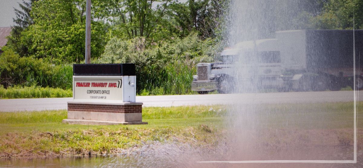 TT in-blog images - TT Blog - 0426-6 Trailer Transit Inc. | A sign for Trailer Transport Inc. stands on a grassy area near a pond, with a road and a driving past —showing that Trailer Transit is who you should hire for power only truck transportation services.