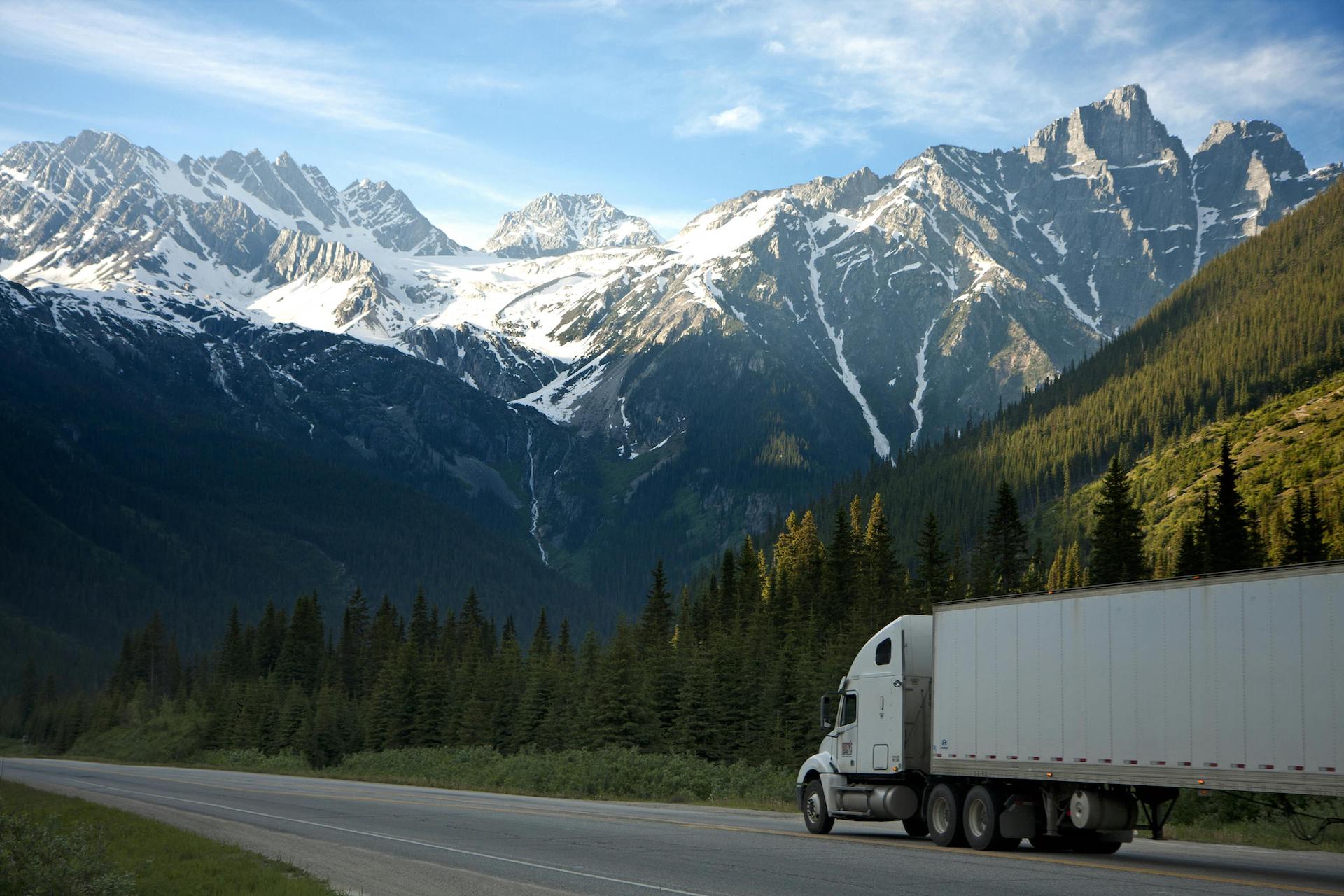 Trailer Transit, Inc. | A white semi-truck drives along a highway with tall pine trees and snow-capped mountains in the background under a partly cloudy sky.