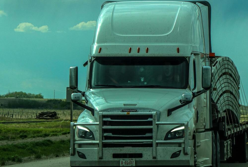 Trailer Transit, Inc. | A white semi-truck with a flatbed trailer carrying large, curved metal pieces drives on a rural highway under a partly cloudy sky.