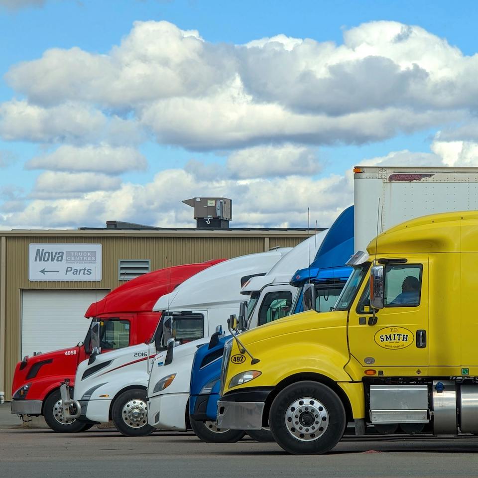Trailer Transit, Inc. | Four semi-trucks in red, white, blue, and yellow are parked side by side outside a truck parts building under a partly cloudy sky.