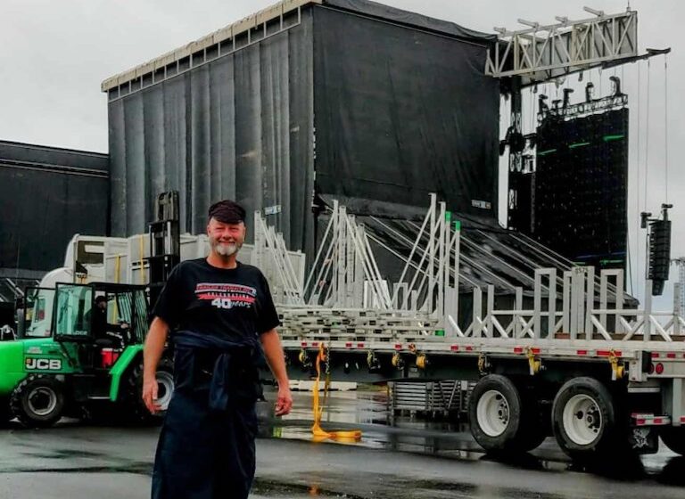 Trailer Transit, Inc. | A man stands in front of a flatbed truck loaded with metal stage equipment, with a forklift and large black stage structures in the background.