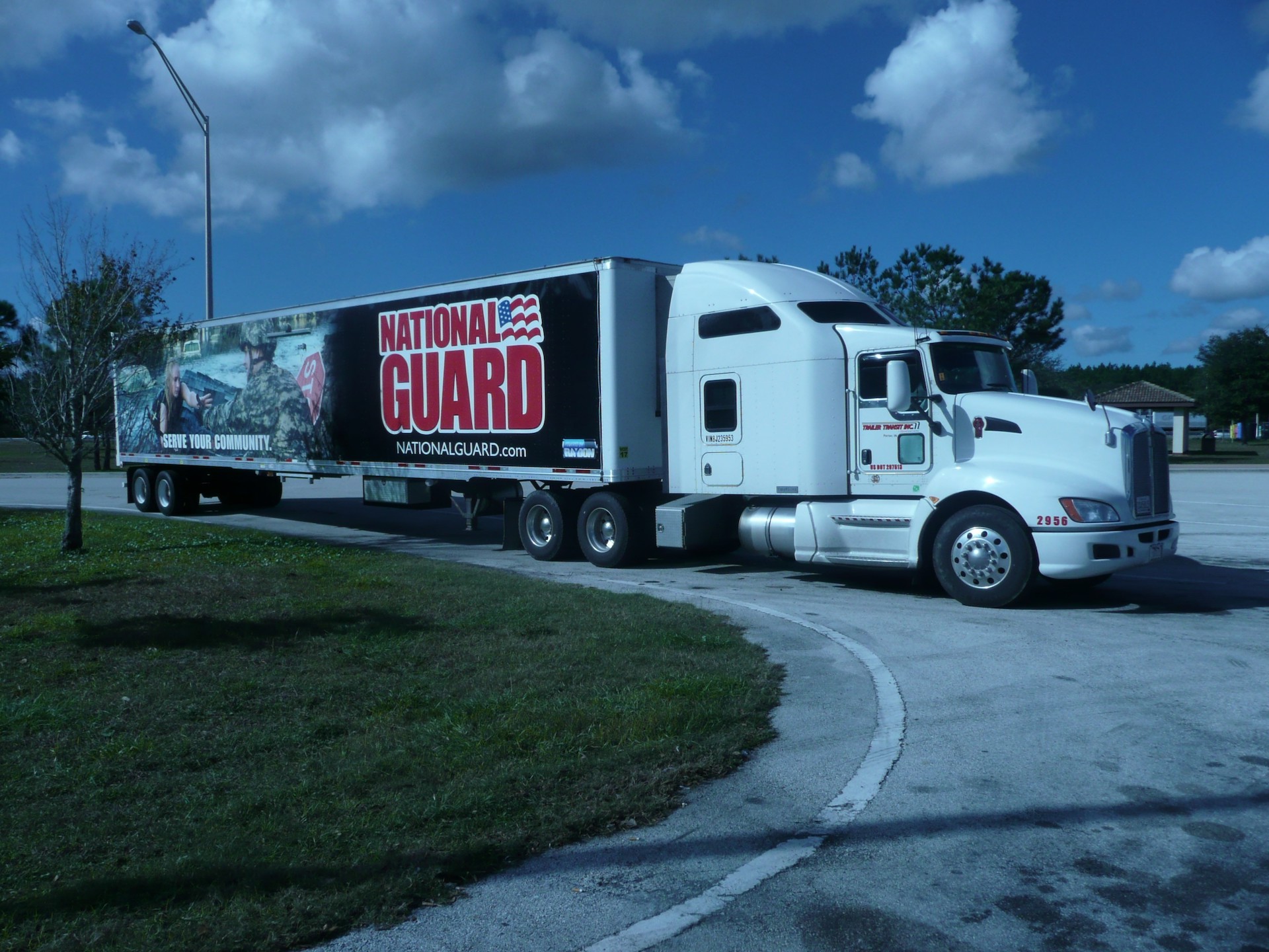 Trailer Transit, Inc. | A white semi-truck with a trailer advertising the National Guard is parked on the side of a road, with trees and a blue sky in the background.