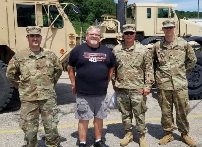 Trailer Transit, Inc. | Four men stand in front of a military truck; three wear camouflage uniforms, and one wears a black T-shirt, gray shorts, and sneakers.