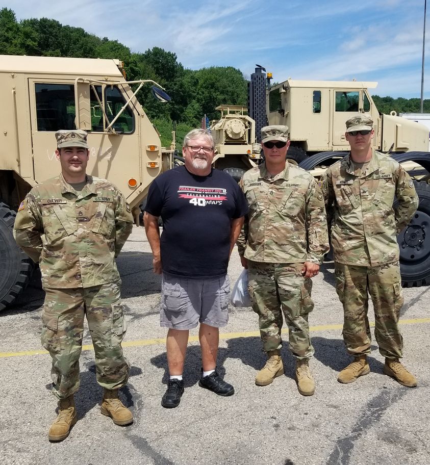 Trailer Transit, Inc. | Four men stand in front of military vehicles; three are in camouflage uniforms, and one is in casual clothes and a t-shirt. The setting appears to be an outdoor parking area.