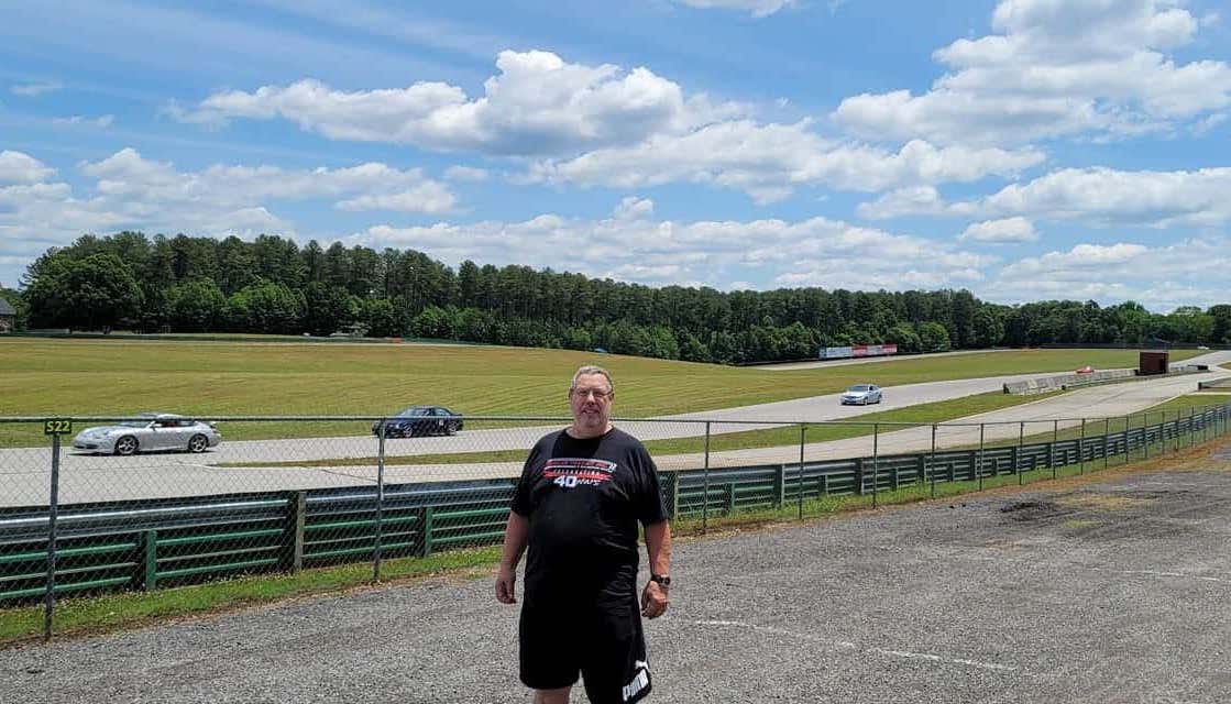 Trailer Transit, Inc. | A man stands in front of a racetrack with several cars driving on it, under a partly cloudy sky with trees and grass in the background.