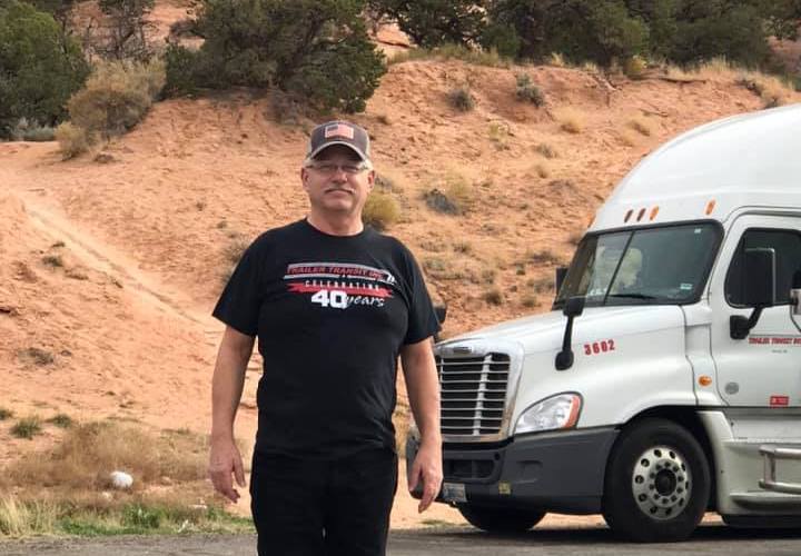 Trailer Transit, Inc. | A man wearing a black t-shirt and cap stands in front of a white semi-truck parked near a desert landscape with shrubs and sand.