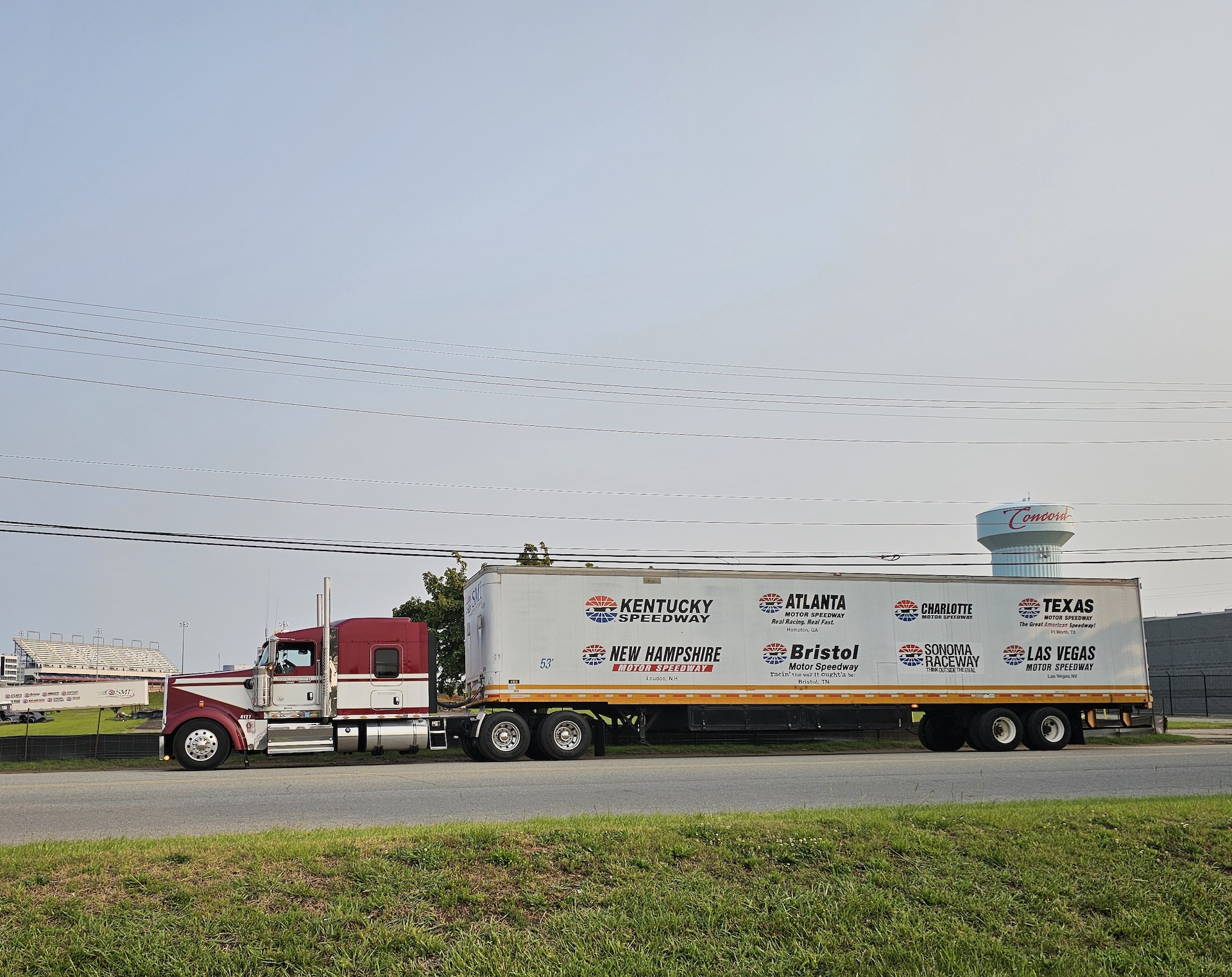 Trailer Transit, Inc. | A semi-truck with a white trailer displaying logos of various speedways is parked on a roadside near a fence and a building.