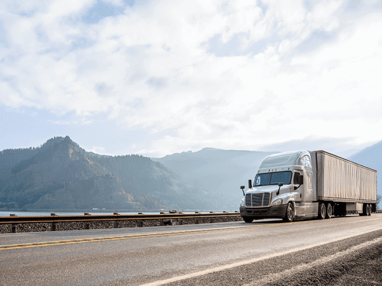 Trailer Transit, Inc. | A semi-truck with a trailer drives on a highway next to a body of water, with mountains and a partly cloudy sky in the background.