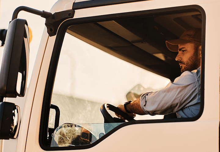 Trailer Transit, Inc. | A man in a cap is sitting in the driver's seat of a truck, looking forward with one hand on the steering wheel.
