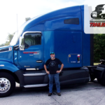 Trailer Transit, Inc. | Owner Operator of the Month, Jeff, stands in front of his blue semi tractor with green trees in the background and a badge at the top with Trailer Transit's Owner Operator of the Month logo.