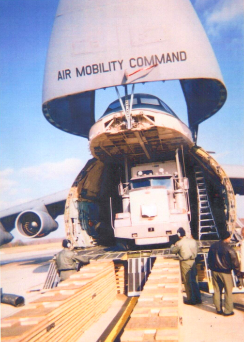 Trailer Transit, Inc. | A military cargo aircraft with its nose open is being loaded with a large truck using wooden ramps, while three people stand nearby.