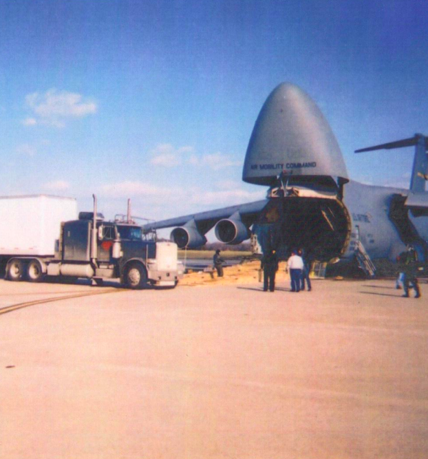 Trailer Transit, Inc. | A large cargo aircraft with its nose open is being loaded with cargo from a semi truck on an airport tarmac, as several people stand nearby.