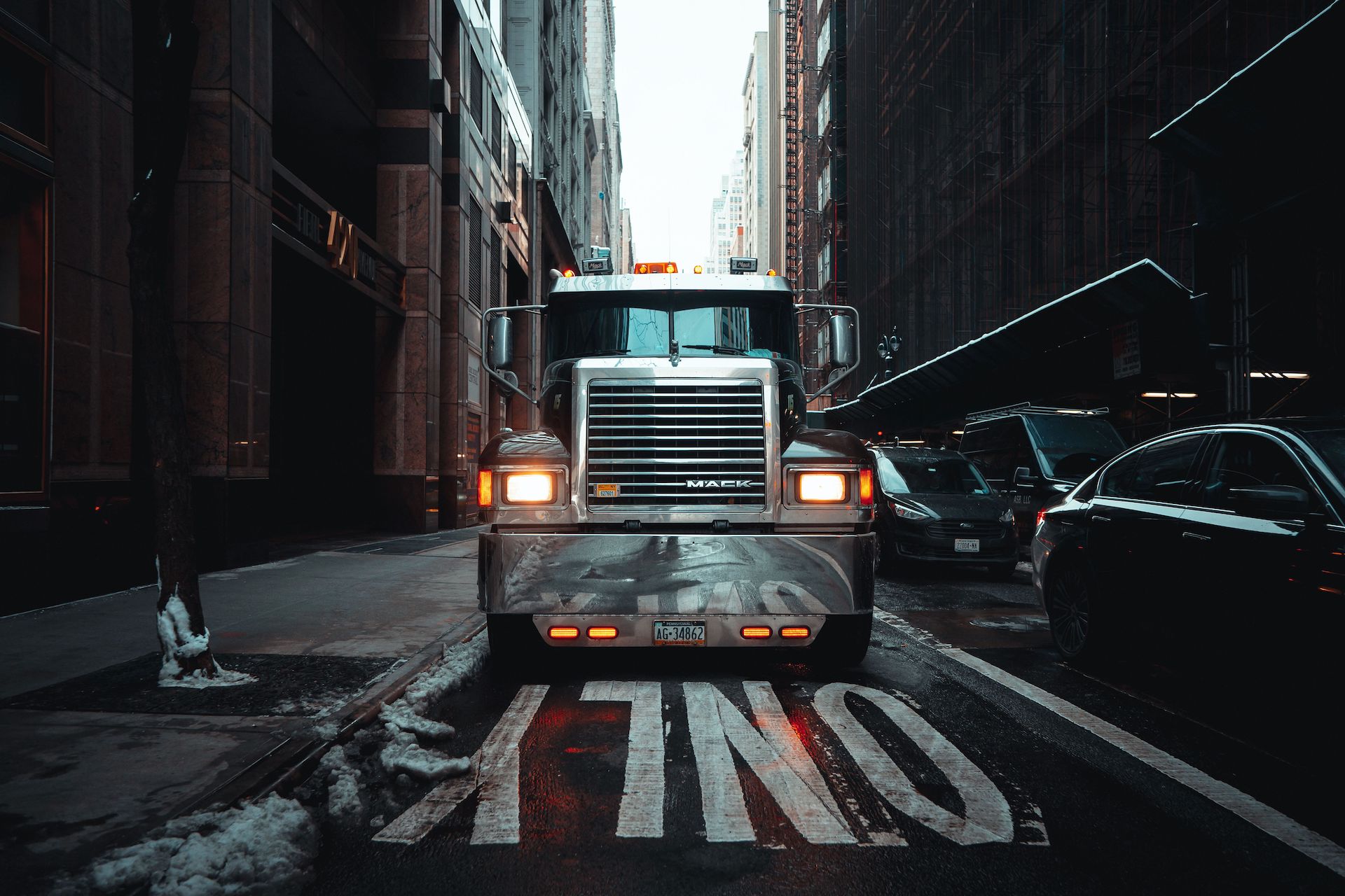 Trailer Transit, Inc. | A large silver Mack truck is parked head-on in the middle of a city street marked "ONLY," surrounded by tall buildings and parked cars.
