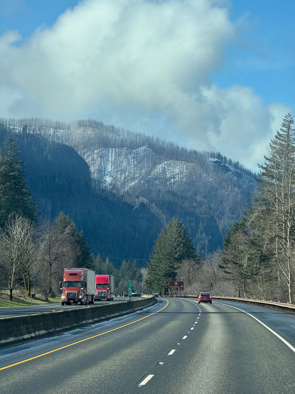 Trailer Transit, Inc. | A highway curves through a forested, mountainous area with two red trucks parked on the side and a red car driving ahead under a partly cloudy sky.