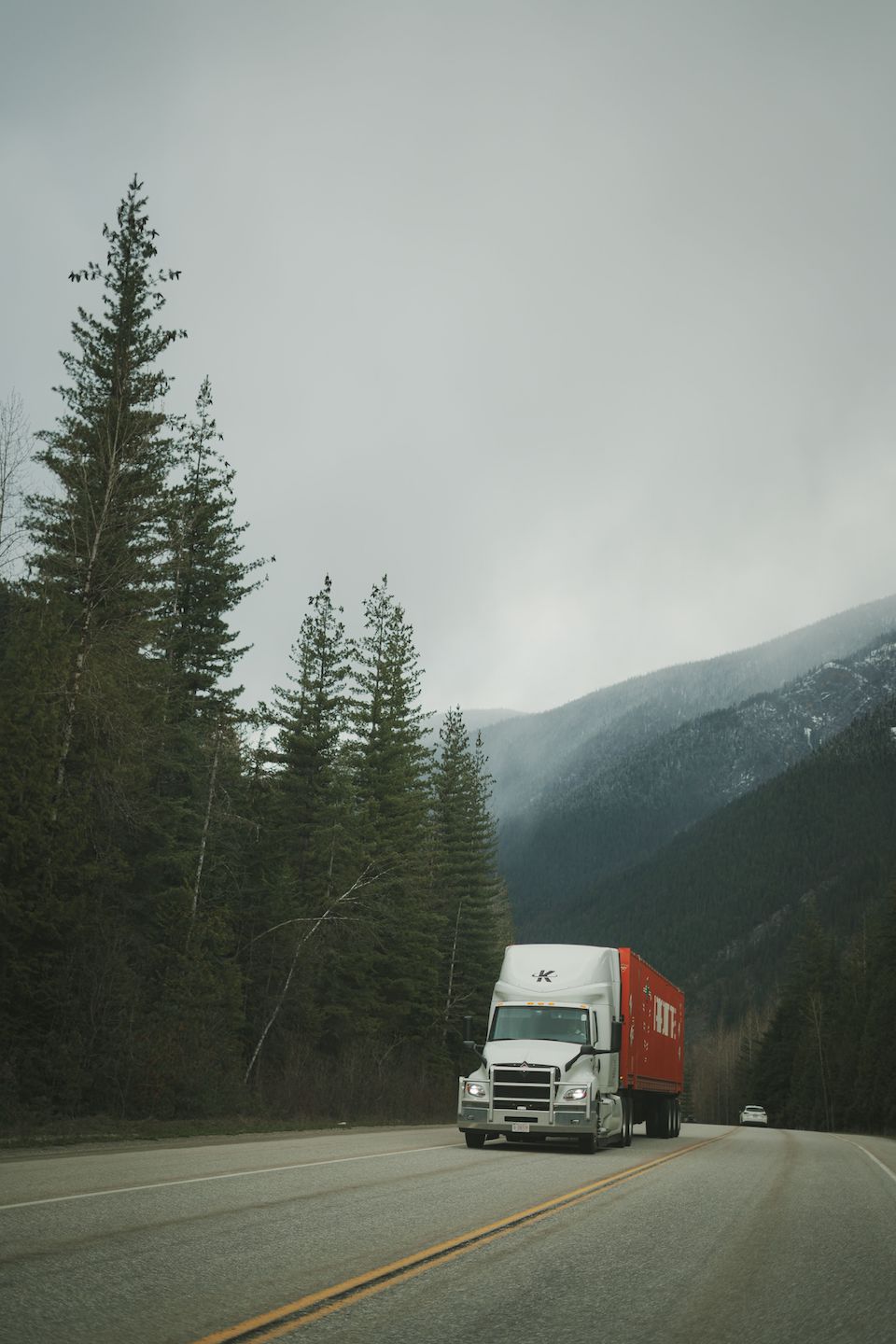 Trailer Transit, Inc. | A white semi-truck with a red container drives on a two-lane road through a forested, mountainous area under a cloudy sky.