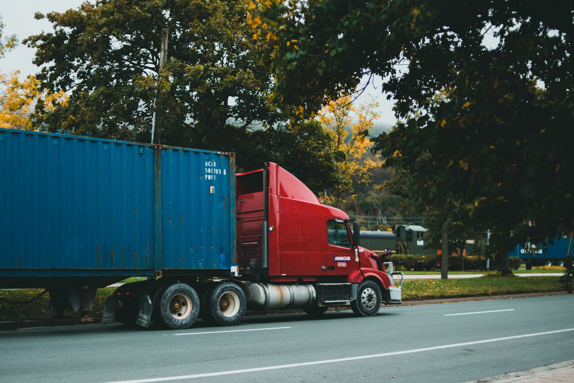 Trailer Transit, Inc. | A red semi-truck with a blue shipping container trailer is parked on the side of a tree-lined street.