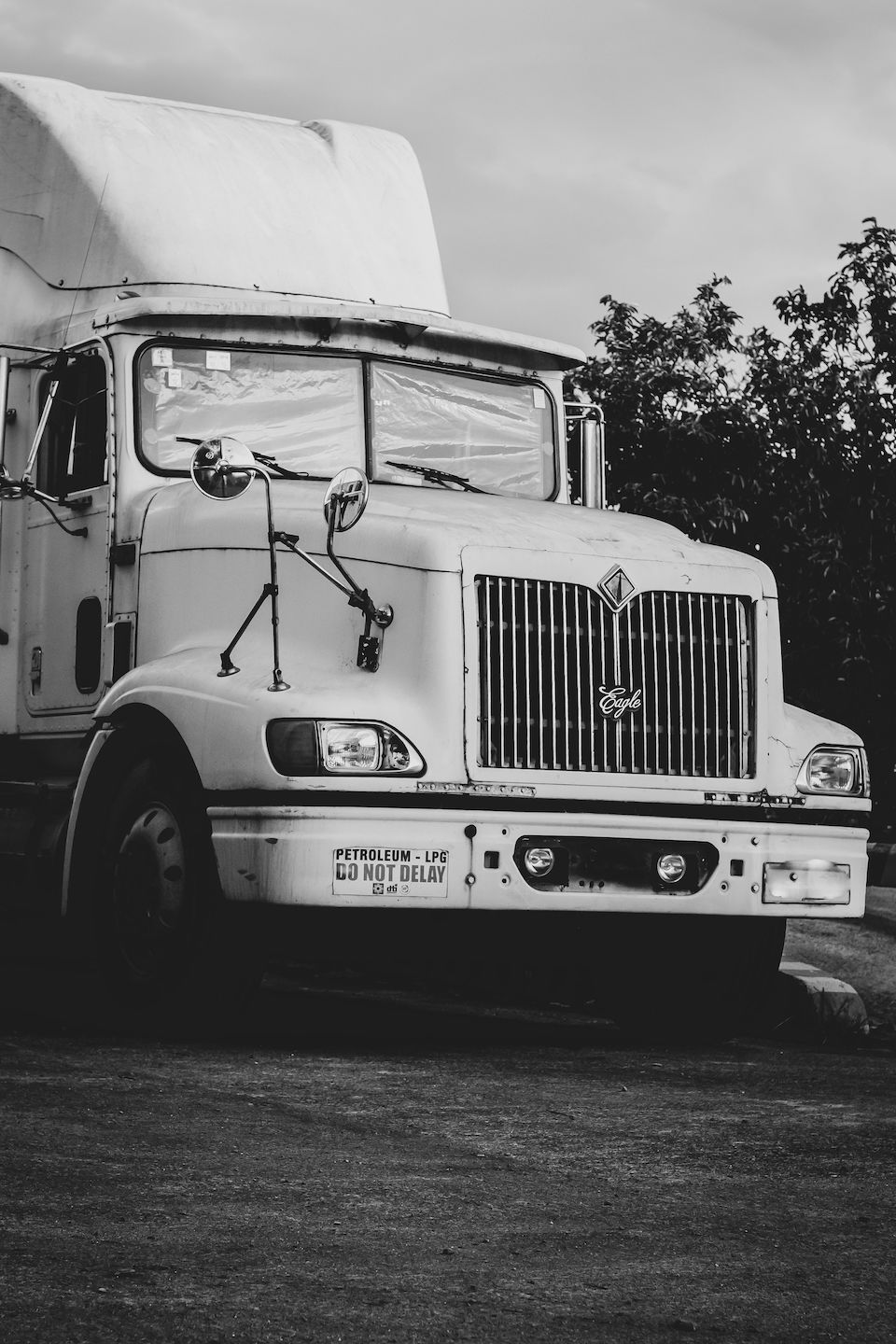 Trailer Transit, Inc. | Black and white photo of a parked semi-truck with a covered cargo area, viewed from the front left side, with trees and cloudy sky in the background.