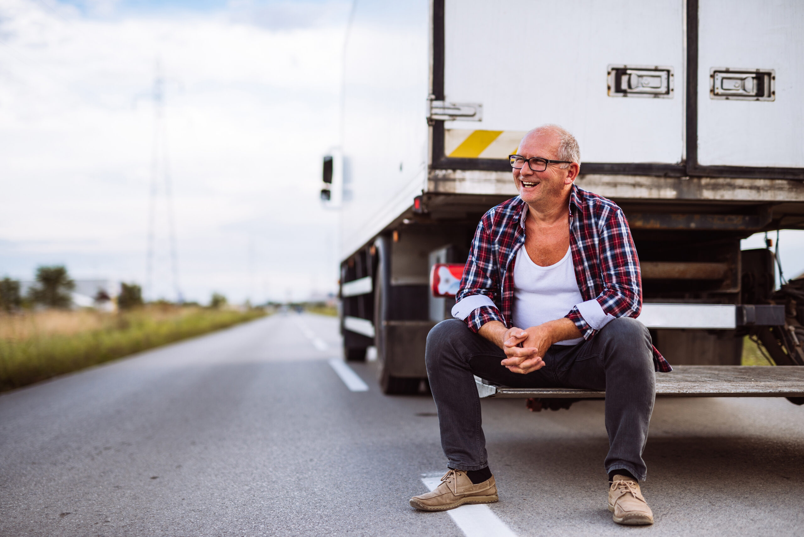 Trailer Transit, Inc. | Smiling man wearing glasses and plaid shirt sits on the open tailgate of a truck parked on the roadside, with fields and a cloudy sky in the background.