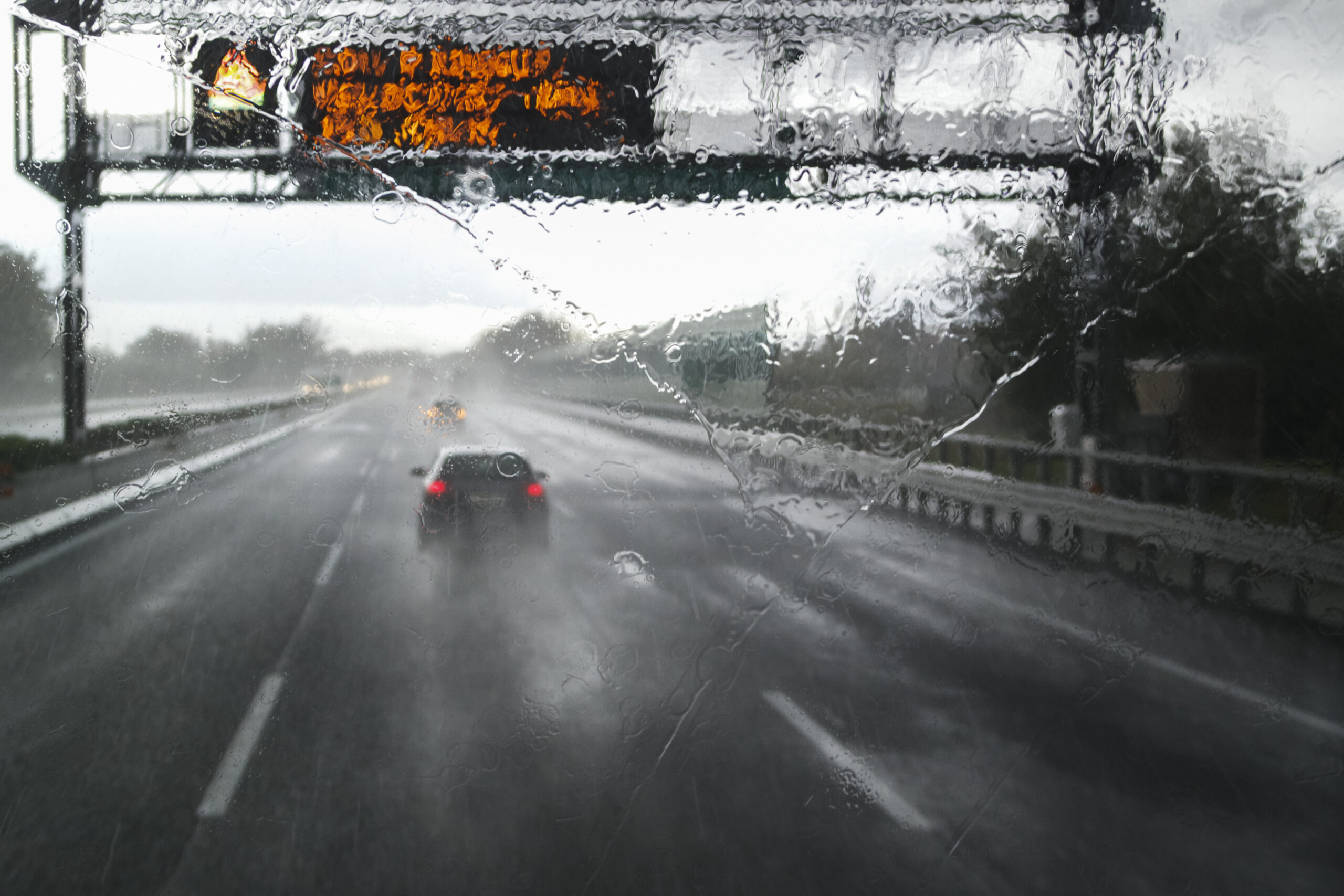Trailer Transit, Inc. | View through a rain-soaked windshield showing cars driving on a wet highway, with an overhead electronic warning sign and blurry surroundings due to heavy rainfall.