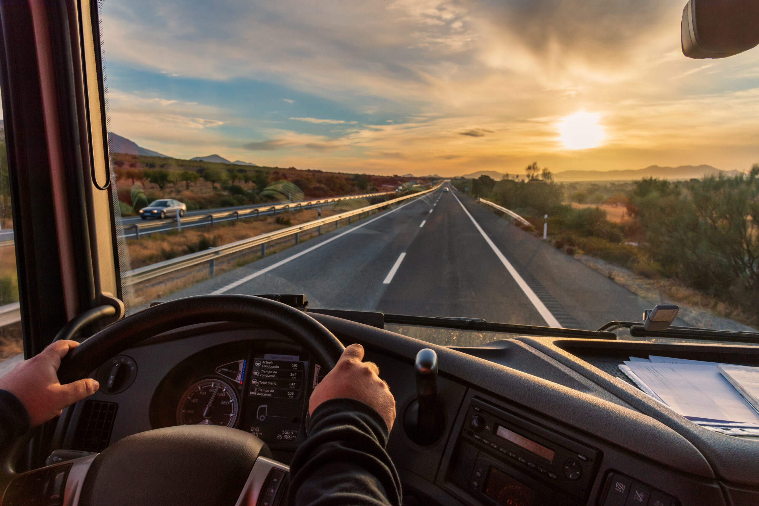 Trailer Transit, Inc. | View from inside a truck cab showing hands on the steering wheel, dashboard instruments, and a highway at sunset with cars driving in the opposite direction.