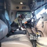 Trailer Transit, Inc. | A dog wearing a vest sits in the driver’s seat of a large truck, facing the steering wheel inside the vehicle’s cab.