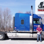 Trailer Transit, Inc. | A man stands next to a blue semi-truck. There are leafless trees in the background and a Trailer Transit Owner Operator of the Month logo in the upper right corner.