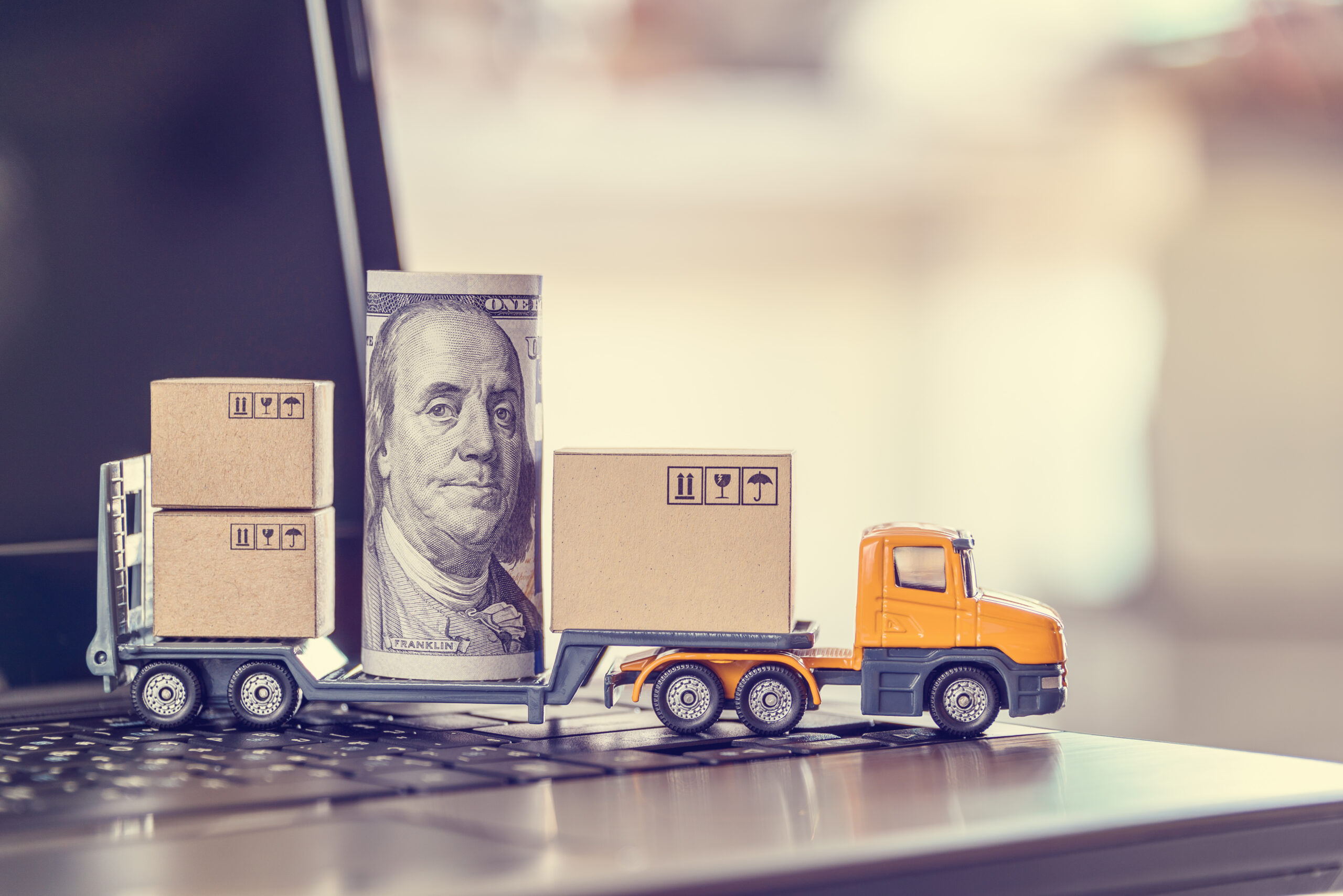 Trailer Transit, Inc. | Toy truck with cardboard boxes and a rolled U.S. hundred dollar bill on a laptop keyboard, representing e-commerce or online shipping and payment.