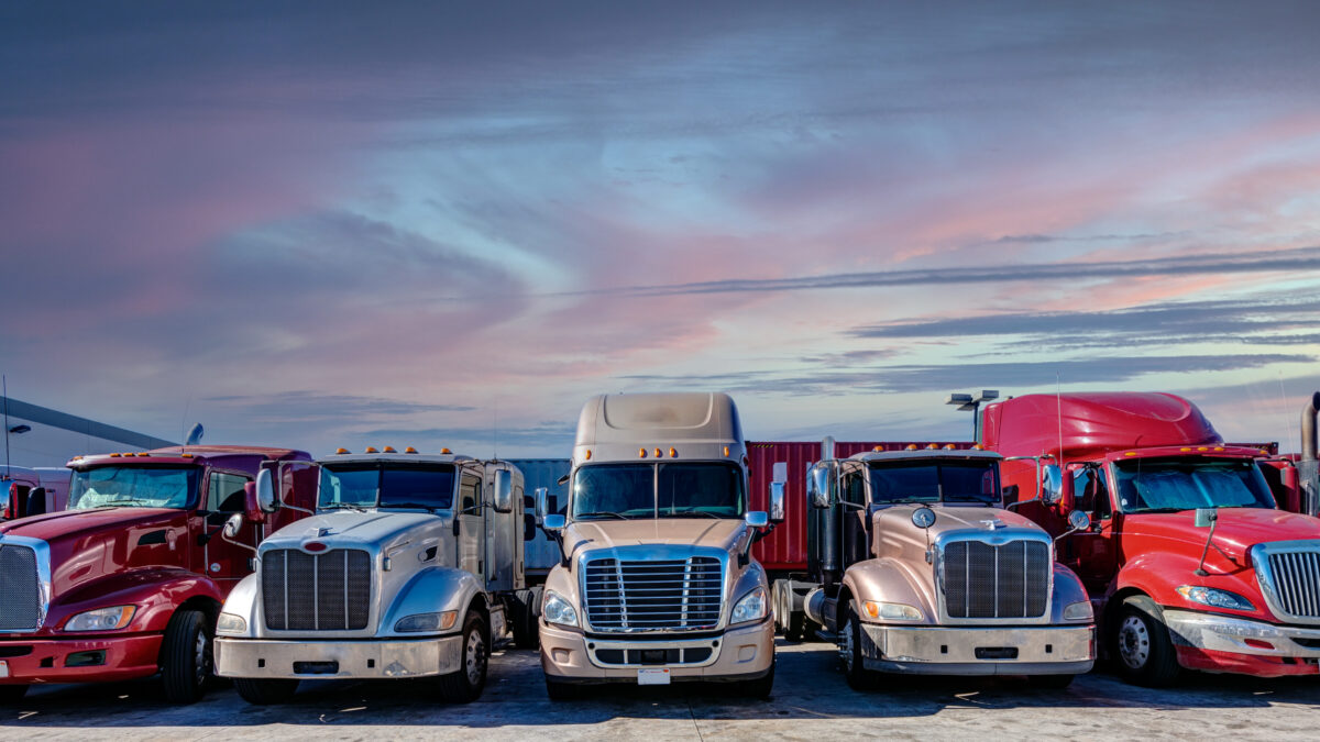 Trailer Transit, Inc. | Six semi-trailer trucks are parked side by side in a lot at sunset, with a pink and blue sky in the background.