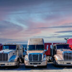 Trailer Transit, Inc. | Six semi-trailer trucks are parked side by side in a lot at sunset, with a pink and blue sky in the background.