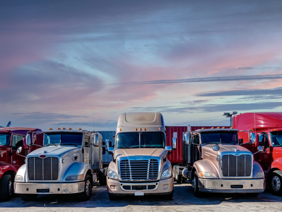 Trailer Transit, Inc. | Six semi-trailer trucks are parked side by side in a lot at sunset, with a pink and blue sky in the background.