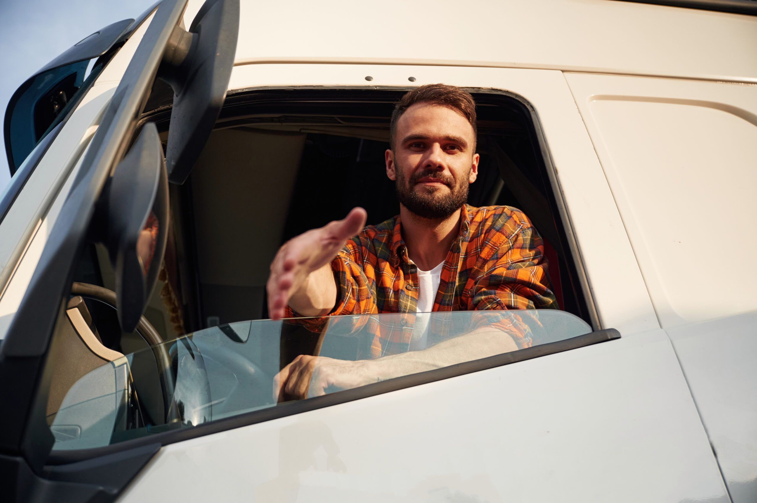 Trailer Transit, Inc. | An Owner Operator in a plaid shirt sits in the driver’s seat of a truck, reaching his hand out of the window as if offering a handshake.