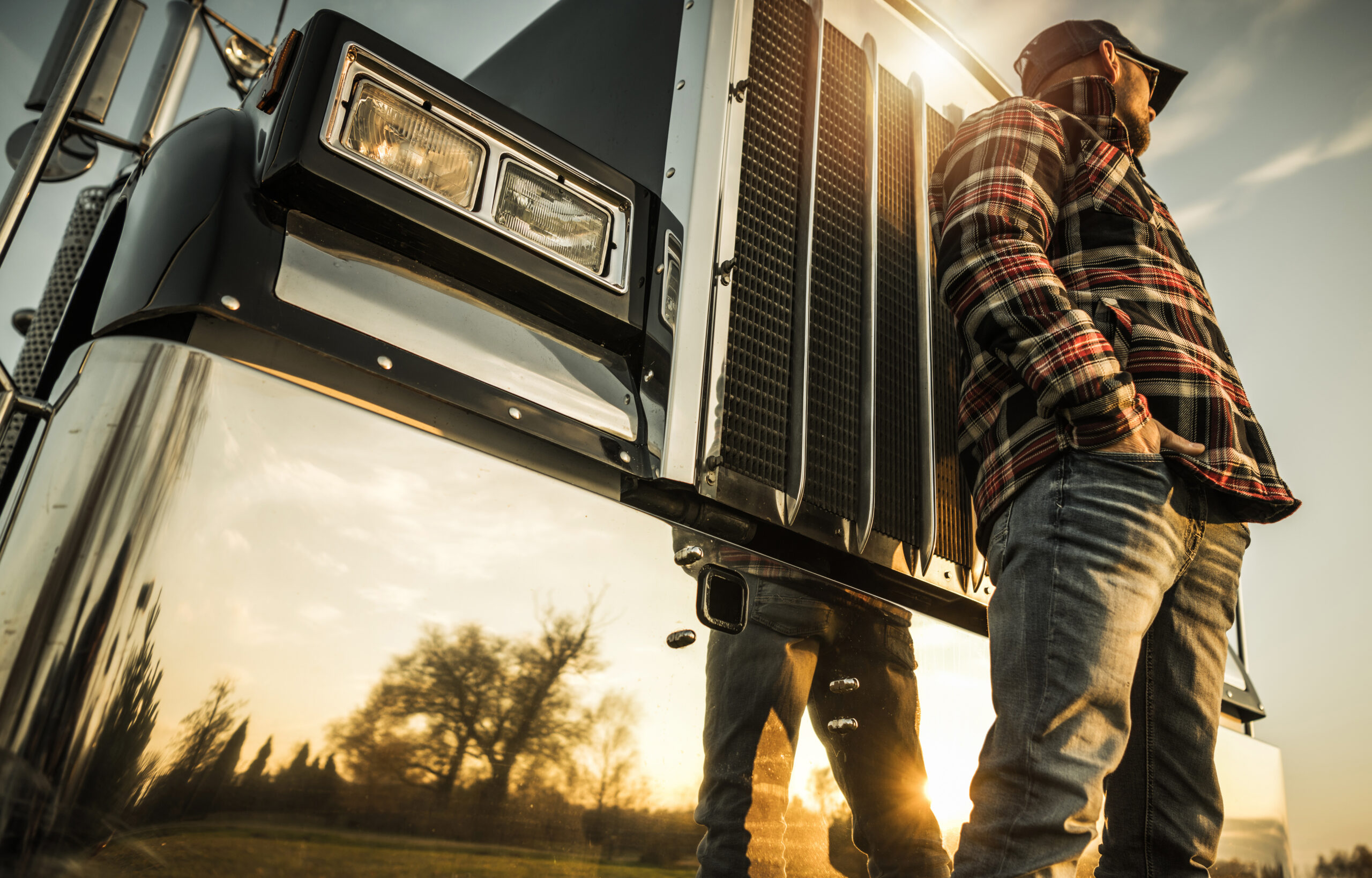 Trailer Transit, Inc. | Owner Operator in jeans and a plaid shirt stands beside a large truck at sunset, with hands in pockets, looking into the distance. The truck’s chrome grille and headlights are visible.
