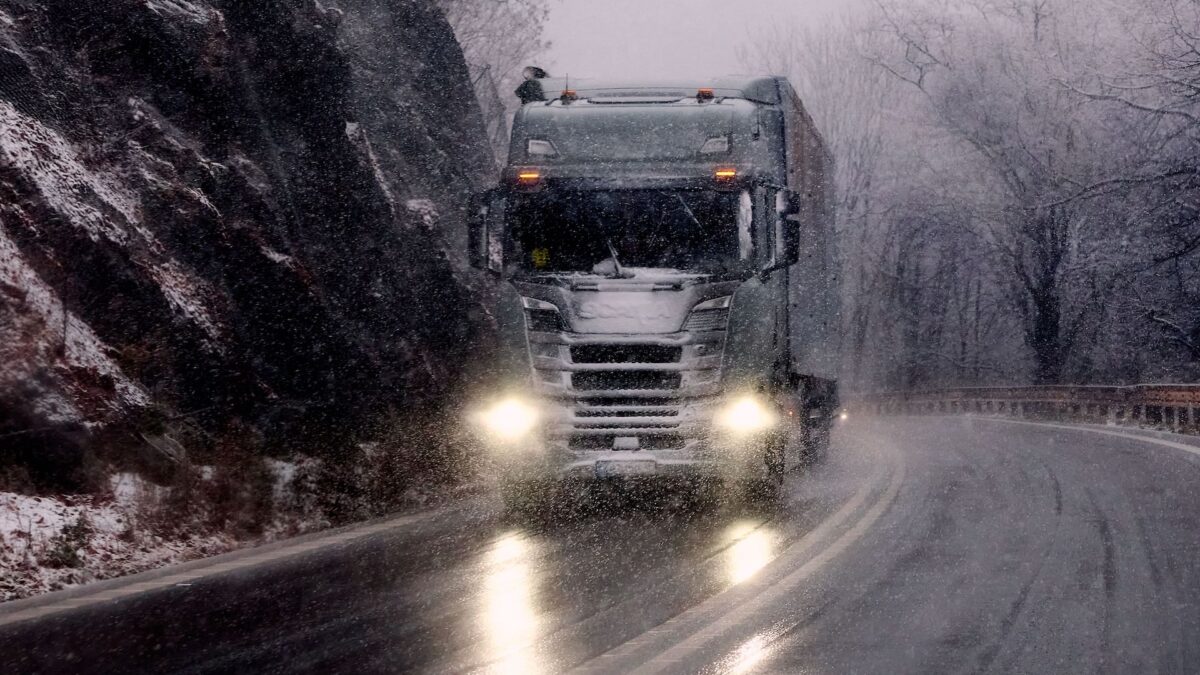 Trailer Transit, Inc. | A semi-truck drives on a winding, snow-covered mountain road in wintry weather, with headlights illuminating the wet pavement.