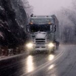 Trailer Transit, Inc. | A semi-truck drives on a winding, snow-covered mountain road in wintry weather, with headlights illuminating the wet pavement.
