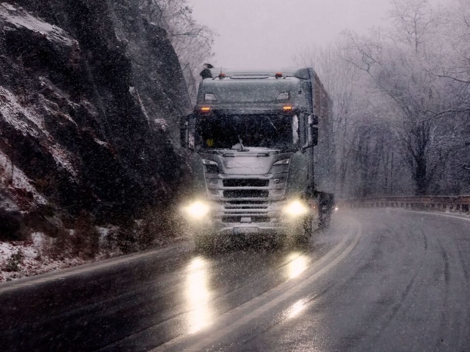 Trailer Transit, Inc. | A semi-truck drives on a winding, snow-covered mountain road in wintry weather, with headlights illuminating the wet pavement.